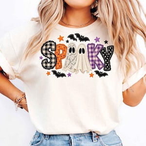 Könnte beinhalten: Ein cremefarbenes T-Shirt mit dem Wort SPOOKY in einem gemusterten Design. Die Buchstaben bestehen aus schwarz-weißem Karomuster, Orange, spitzenartigen Texturen und violettem Gingham. Zwei weiße Geister und schwarze Fledermäuse sind abgebildet.