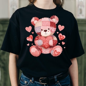 Könnte beinhalten: Schwarzes T-Shirt mit einem Patchwork-Teddybären, der ein Herz hält. Der Bär besteht aus rosa, roten und lila gemusterten Stoffen. Rosa Herzen sind um den Bären herum verstreut. Das T-Shirt hat einen lässigen Stil.