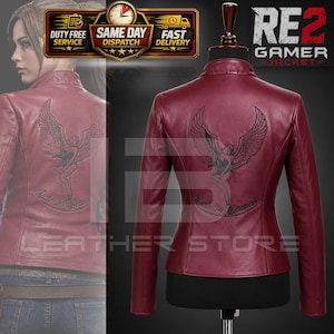 May include: A maroon leather jacket featuring a stand-up collar and long sleeves. The back displays a black embroidered graphic of a winged figure with the text Made In Heaven. The jacket is displayed on a mannequin and also shown on a person.