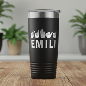 May include: A black travel tumbler with a silver lid. The tumbler features the name "EMILI" in white, spelled out using American Sign Language handshapes. The tumbler is cylindrical with a ridged base.