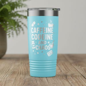 Puede incluir: Un vaso térmico azul claro con tapa plateada. El vaso presenta las palabras "Caffeine, Codeine, and Chaos" en letras plateadas, junto con iconos relacionados como una taza de café y pastillas. El vaso es cilíndrico con una base acanalada.