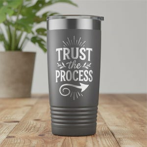 May include: A dark gray travel tumbler with a silver lid. The tumbler features the white text "TRUST the PROCESS" with an arrow graphic. The tumbler is cylindrical with horizontal ridges near the bottom.