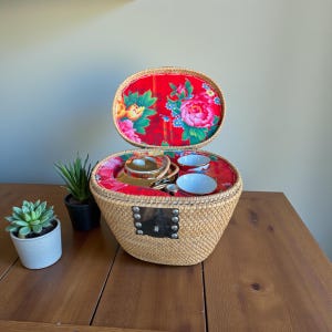 Könnte beinhalten: Ein Vintage-Teeservice in einem geflochtenen Korb. Der Korb hat ein rot geblümtes Futter und einen Deckel. Das Set enthält eine goldene Teekanne, zwei kleine weiße Tassen und einen kleinen Behälter. Der Korb steht auf einem Holztisch.