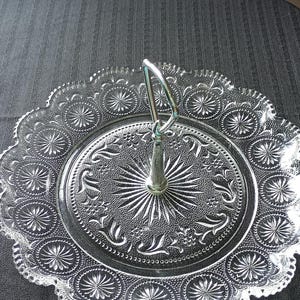 May include: A clear pressed glass serving tray featuring a central metal handle. The circular plate has a scalloped edge and is decorated with intricate embossed starburst patterns and floral motifs throughout the surface of the glass.