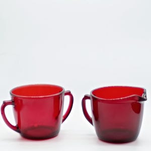 May include: A pair of translucent ruby red glass vessels, consisting of a small handled mug and a matching creamer pitcher with a pouring spout. Both items feature a smooth, rounded design and a deep, consistent red color throughout the glass.