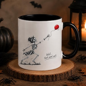 May include: A white ceramic mug with a black interior and handle, featuring a black and white illustration of a skeleton running while reaching for a red balloon. The text "Well... that went well." is printed in black ink below the balloon.