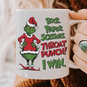 May include: White ceramic mug with a green cartoon character in a red Santa suit. The mug has the text "Rock, Paper, Scissors, Throat Punch! I Win." The character is wearing a Santa hat. The mug is held by a person.