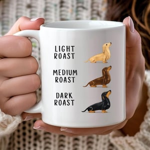 May include: White ceramic coffee mug with illustrations of dachshunds representing coffee roasts. The mug has the words "LIGHT ROAST", "MEDIUM ROAST", and "DARK ROAST" printed in black. The mug is held by a person.