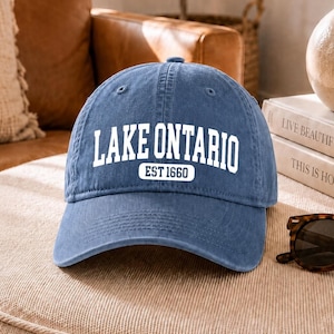 May include: A blue baseball cap with a curved brim and a six-panel construction. The front features white embroidered text that reads LAKE ONTARIO with the text EST 1660 enclosed in a white rectangular border directly underneath the main lettering.