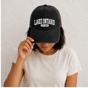 May include: A charcoal black baseball cap with a distressed, washed-out texture. The front of the cap features white embroidered text that reads LAKE ONTARIO in a bold, arched font, with the smaller text EST 1660 centered directly underneath.