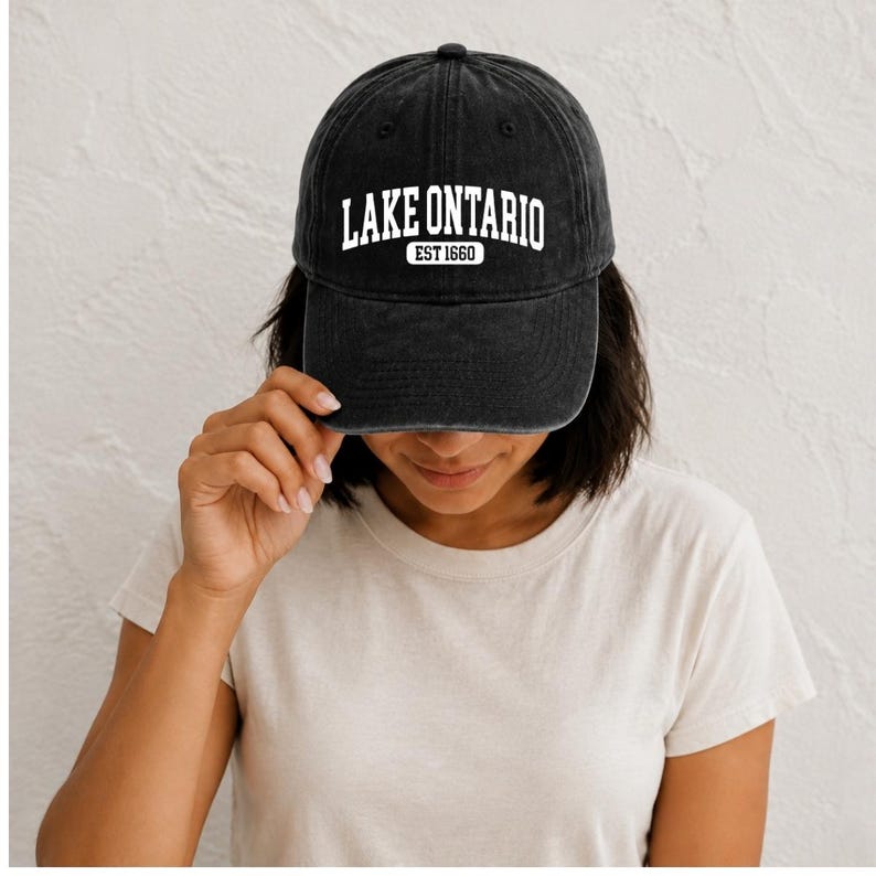 May include: A charcoal black baseball cap with a distressed, washed-out texture. The front of the cap features white embroidered text that reads LAKE ONTARIO in a bold, arched font, with the smaller text EST 1660 centered directly underneath.