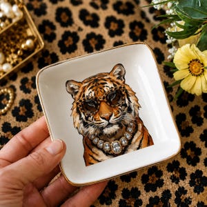 May include: A square white ceramic trinket dish with a gold-toned rim. The center features a detailed illustration of a tiger wearing leopard-print sunglasses and a chunky, sparkling crystal-style necklace against a clean white background.