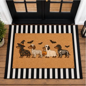Puede incluir: Un felpudo rectangular de fibra de coco color canela con cuatro perros salchicha disfrazados de Halloween: una bruja, una momia, un fantasma con un cubo de calabaza y un esqueleto. Murci&eacute;lagos negros aparecen ilustrados sobre los perros.