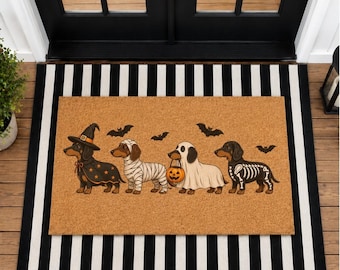 Könnte beinhalten: Eine rechteckige, hellbraune Kokosfasermatte mit vier Dackeln in Halloween-Kostümen: eine Hexe, eine Mumie, ein Geist mit Kürbiseimer und ein Skelett. Über den Hunden sind schwarze Fledermäuse auf die Matte gedruckt.