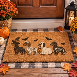 Puede incluir: Un felpudo rectangular de fibra de coco color canela con cuatro perros salchicha disfrazados de Halloween: una bruja, una momia, un fantasma con una calabaza y un esqueleto. Siluetas de murci&eacute;lagos negros aparecen dispersas sobre los perros.