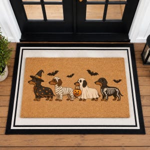 Puede incluir: Un felpudo rectangular de fibra de coco color canela con cuatro perros salchicha disfrazados de Halloween: una bruja, una momia, un fantasma con un cubo de calabaza y un esqueleto. Murci&eacute;lagos negros est&aacute;n impresos sobre los perros.