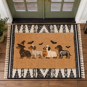 Puede incluir: Un felpudo rectangular de fibra de coco color canela con cuatro perros salchicha disfrazados de Halloween: bruja, momia, fantasma con un cubo de calabaza y esqueleto. Murci&eacute;lagos negros est&aacute;n impresos sobre los perros. Descansa sobre una alfombra blanca y negra.