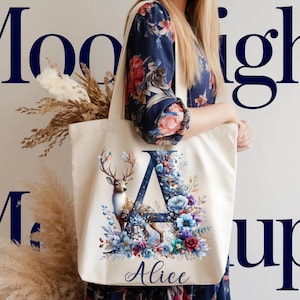 May include: Cream-colored tote bag featuring a deer illustration with floral accents and the letter "A". The design includes blue, purple, and red flowers, with the name "Alice" printed below. The bag is held by a person.