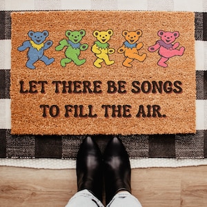 May include: A rectangular doormat with a brown surface features five colorful dancing bear illustrations. The text "LET THERE BE SONGS TO FILL THE AIR" is printed in brown. Black boots and light blue jeans are visible.