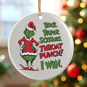 May include: A circular Christmas decoration with a Grinch character in a red Santa suit and hat. The ornament displays the text "Rock, Paper, Scissors, Throat Punch! I Win." in green and red. A gold string is attached for hanging.