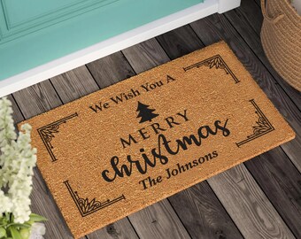 May include: A rectangular doormat made of natural fibers. The mat features the text "We Wish You A Merry Christmas" with a Christmas tree graphic and the family name "The Johnsons" in black lettering.