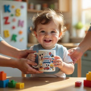 K&ouml;nnte beinhalten: Ein l&auml;chelndes Baby h&auml;lt einen wei&szlig;en Becher mit Henkel. Der Becher ist mit Zeichentrickfiguren, Feuerwerk und dem Text "Little American Cutie 1776-2022" verziert. Das Baby tr&auml;gt eine blaue L&auml;tzchen und ein wei&szlig;es T-Shirt.