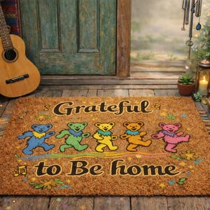 May include: A rectangular brown doormat with the words "Grateful to Be Home" and dancing bear graphics in blue, green, yellow, orange, and pink. The mat is placed in front of a green door and a wooden guitar.
