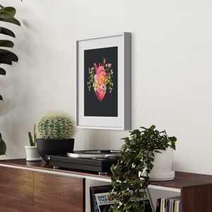 May include: A framed print of a red anatomical heart with pink and yellow flowers growing from it, on a black background. The frame is white and the print is hanging on a white wall.