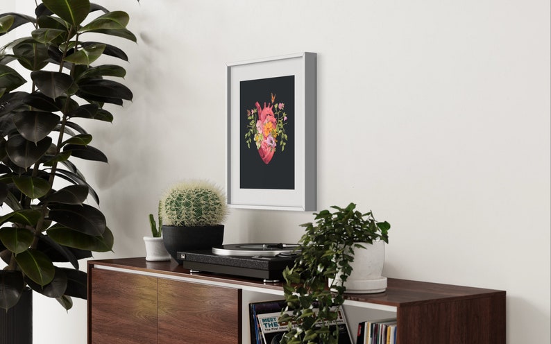 May include: A framed print of a red anatomical heart with pink and yellow flowers growing from it, on a black background. The frame is white and the print is hanging on a white wall.