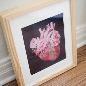 May include: A framed print of a realistic anatomical heart with pink roses growing from it. The heart is painted in shades of pink and red, and the roses are a light pink. The background is a dark brown.