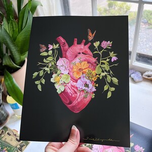 May include: A black and white print of a human heart with pink and red tones, surrounded by a variety of flowers and butterflies in shades of pink, yellow, and green. The print is signed in gold lettering.