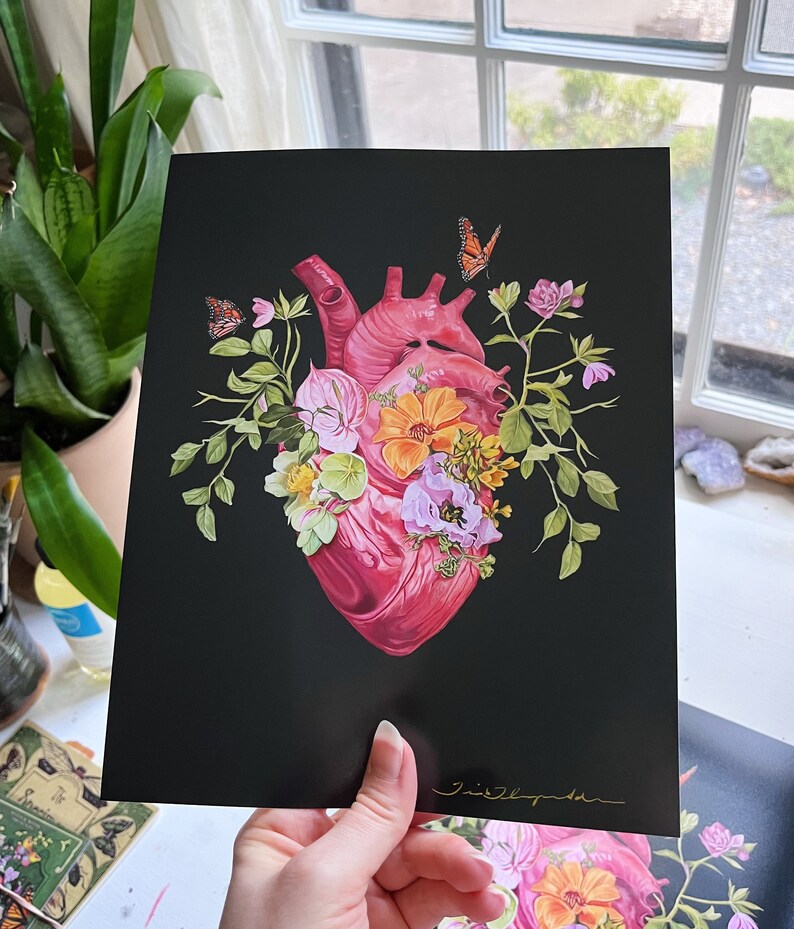 May include: A black and white print of a human heart with pink and red tones, surrounded by a variety of flowers and butterflies in shades of pink, yellow, and green. The print is signed in gold lettering.