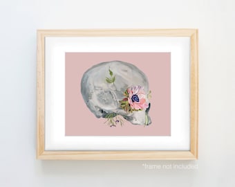 Può includere: Un teschio umano bianco con un fiore rosa e foglie verdi che crescono dalla cavità oculare, su uno sfondo rosa.