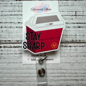 May include: A red and white badge reel featuring a sharps container design with the words "STAY SHARP". The badge reel has a clear plastic strap and a metal snap closure. The background includes a white card with the words "Heavenly Stars".