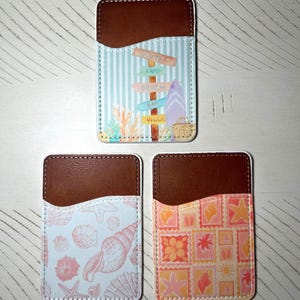 May include: Three phone card holders with brown leather tops and white stitching. One has a beach-themed design with a signpost, surfboard, and text. The other two have seashell and stamp designs.