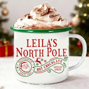 Peut inclure: Mug en émail blanc avec un bord vert, rempli de chocolat chaud et de crème fouettée, saupoudré de cacao. Le mug indique "LEILA'S NORTH POLE AUTHENTIC HOT CHOCOLATE SINCE 1897 SANTA CLAUS APPROVED". Thème de Noël.