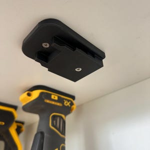 May include: A black 3D-printed power tool battery holder, mounted with screws. The holder is rectangular with a slot for a battery and is attached to a white surface. A yellow and black power tool is visible below.