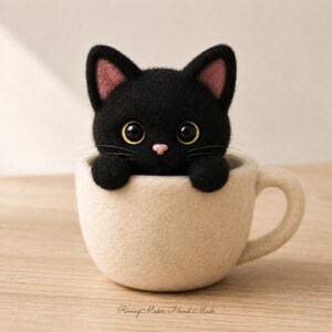 Può includere: Una statuetta di gatto nero in feltro ad ago con grandi occhi scuri e un piccolo naso rosa, posizionata all'interno di una tazza in feltro color crema. Il gatto ha orecchie a punta con dettagli rosa e baffi. Testo Roving Maker Hand Made.