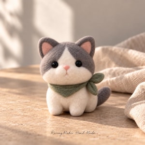 Puede incluir: Una figura de gato de lana fieltrada con pelaje gris y blanco, ojos negros, nariz rosa y orejas rosadas por dentro. El gato lleva una bufanda verde salvia atada al cuello. El texto Roving Maker Hand Made está impreso en la parte inferior.