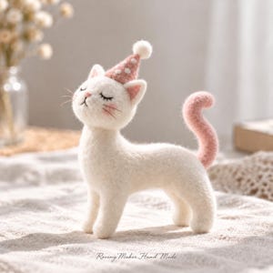 Könnte beinhalten: Eine nadelgefilzte weiße Katzenfigur mit einem rosa Partyhut mit weißen Punkten und einem Bommel. Die Katze hat geschlossene Augen, rosa Wangen und einen geschwungenen rosa Schwanz. Der Text Roving Maker Hand Made steht unten.