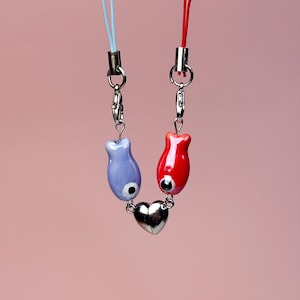 May include: A necklace with two fish-shaped charms, one red and one blue, linked by a silver heart pendant. The charms have black eye details and hang from a blue and red cord.