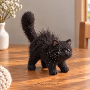 Pode incluir: Uma estatueta de gato preto feita em feltro de agulha com textura felpuda, cauda erguida e olhos grandes e escuros. O gato tem longos bigodes brancos e pequenas orelhas com bordas cor-de-rosa. O texto Roving Maker Hand Made está impresso.