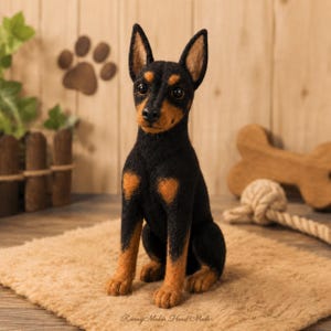 May include: A needle-felted miniature pinscher dog figurine with black and tan fur, pointed ears, and dark eyes. The sculpture is in a seated position, featuring a textured wool finish. The text Roving Maker Hand Made is visible at the bottom.