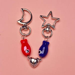 May include: A keychain featuring a silver-tone clasp in the shape of a crescent moon and a star. The keychain has a red and a blue bead with an eye design, connected by a silver heart charm.
