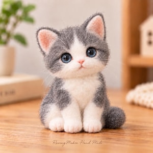 May include: A needle-felted kitten figurine with grey and white fur, large blue eyes, and pink inner ears. The cat is sitting in an upright position with a white chest and paws. The text Roving Maker Hand Made is printed at the bottom centre.