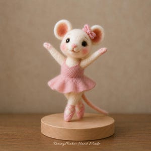 May include: A needle felted mouse figurine wearing a pink tutu and matching ballet slippers. The white mouse has pink inner ears, a small pink bow on its head, and rosy cheeks. It stands on a round wooden base with the text RovingMaker Hand Made.