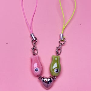 May include: A pink and green phone charm with a silver heart-shaped clasp. The charm features two ceramic fish-shaped beads with googly eyes, connected by a silver chain. The pink cord is 6 inches long, and the green cord is 6 inches long.