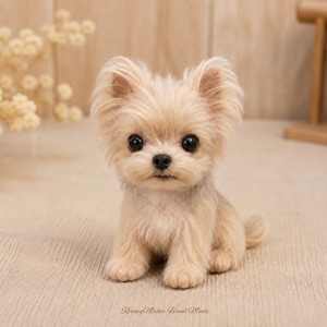 May include: A small, needle-felted figurine of a puppy with light beige, fluffy fur, upright ears, and large, round, black eyes. The puppy is sitting in an alert posture, featuring a small black nose and a textured, soft appearance.