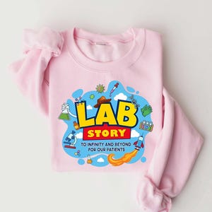 May include: A light pink sweatshirt with the words "LAB STORY" in yellow and red, with a science-themed graphic. The graphic includes a microscope, test tubes, a rocket, and the text "TO INFINITY AND BEYOND FOR OUR PATIENTS."