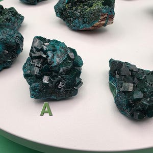 May include: A collection of raw, teal-colored mineral specimens with crystalline structures. The minerals are displayed on a white, circular platform. The letter "A" is visible on the platform. The text "NMOIIOMN 4 GalaxyXGems" is at the bottom.